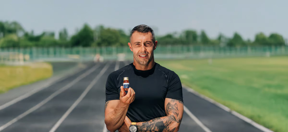 The smiling Ironman record holder Jurand Czabański stands on the running track, holding a bottle of Primabiotic Collagen Sport in his hand