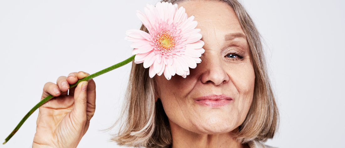 Smiling woman during menopause holding a pink flower over one eye, showing off her radiant skin and beautiful hair after taking collagen shots