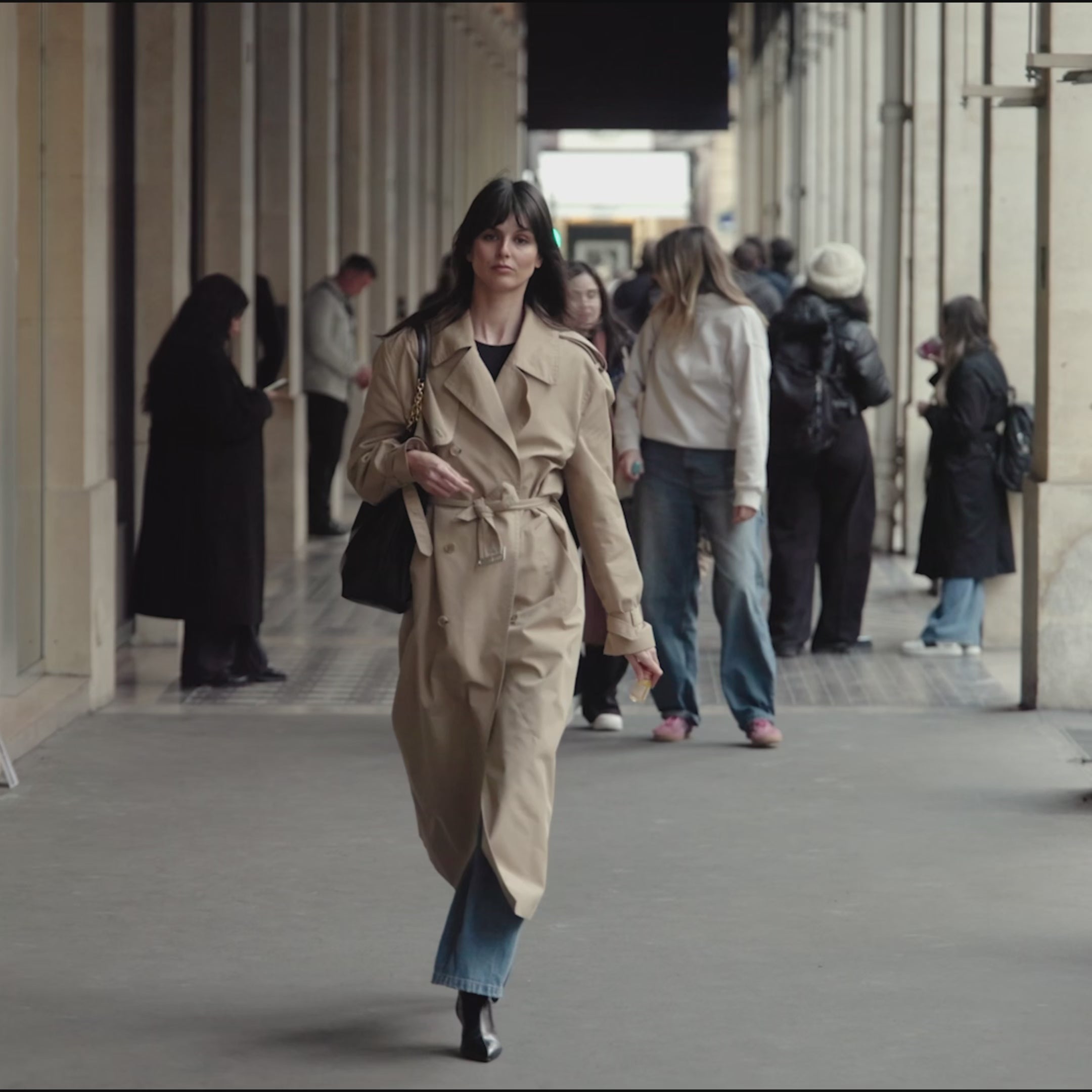 Stylish woman in a beige trench coat confidently walking through a city arcade, embodying natural beauty and elegance