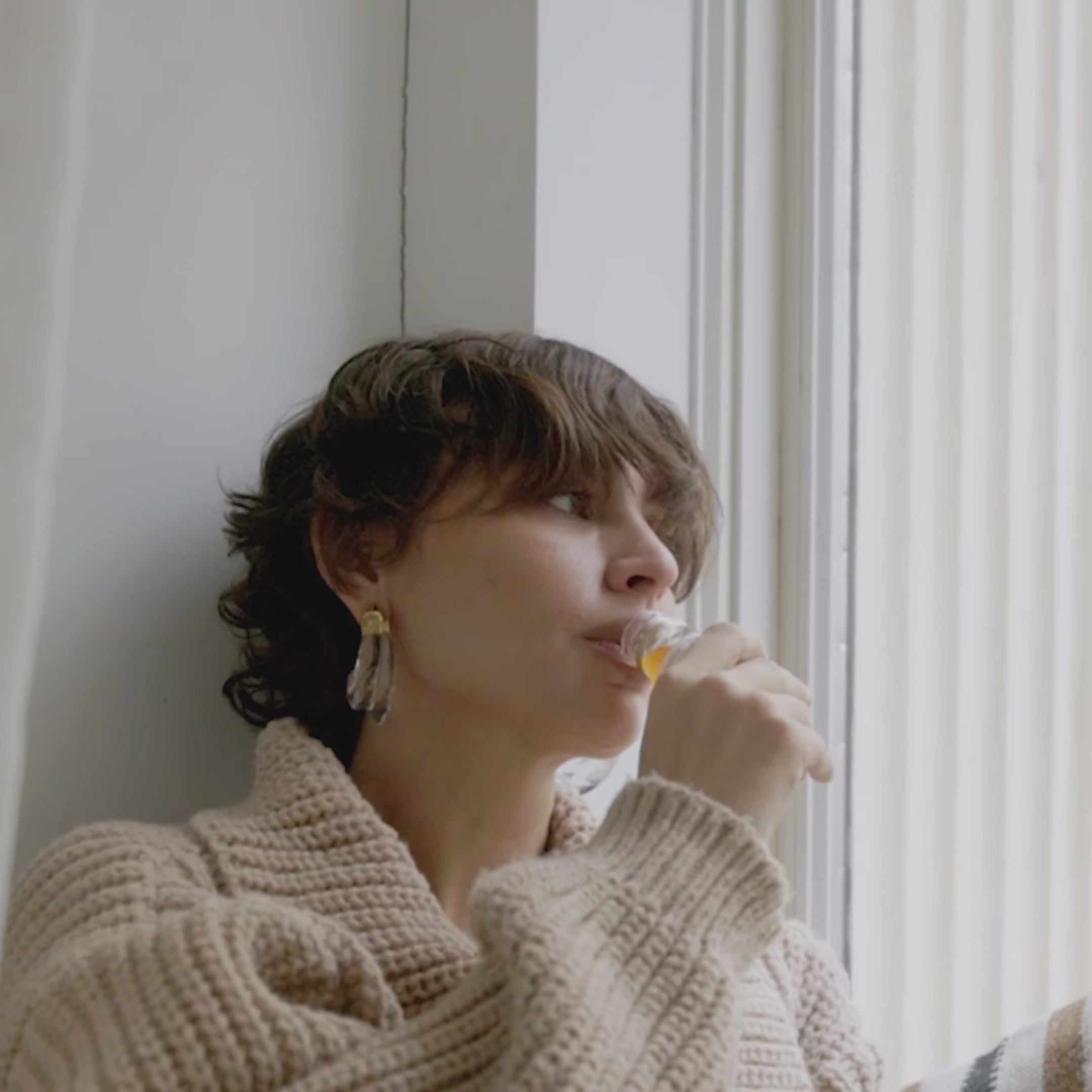 Woman in a cosy sweater drinks Primabiotic Collagen by the window, enjoying a relaxing beauty ritual.