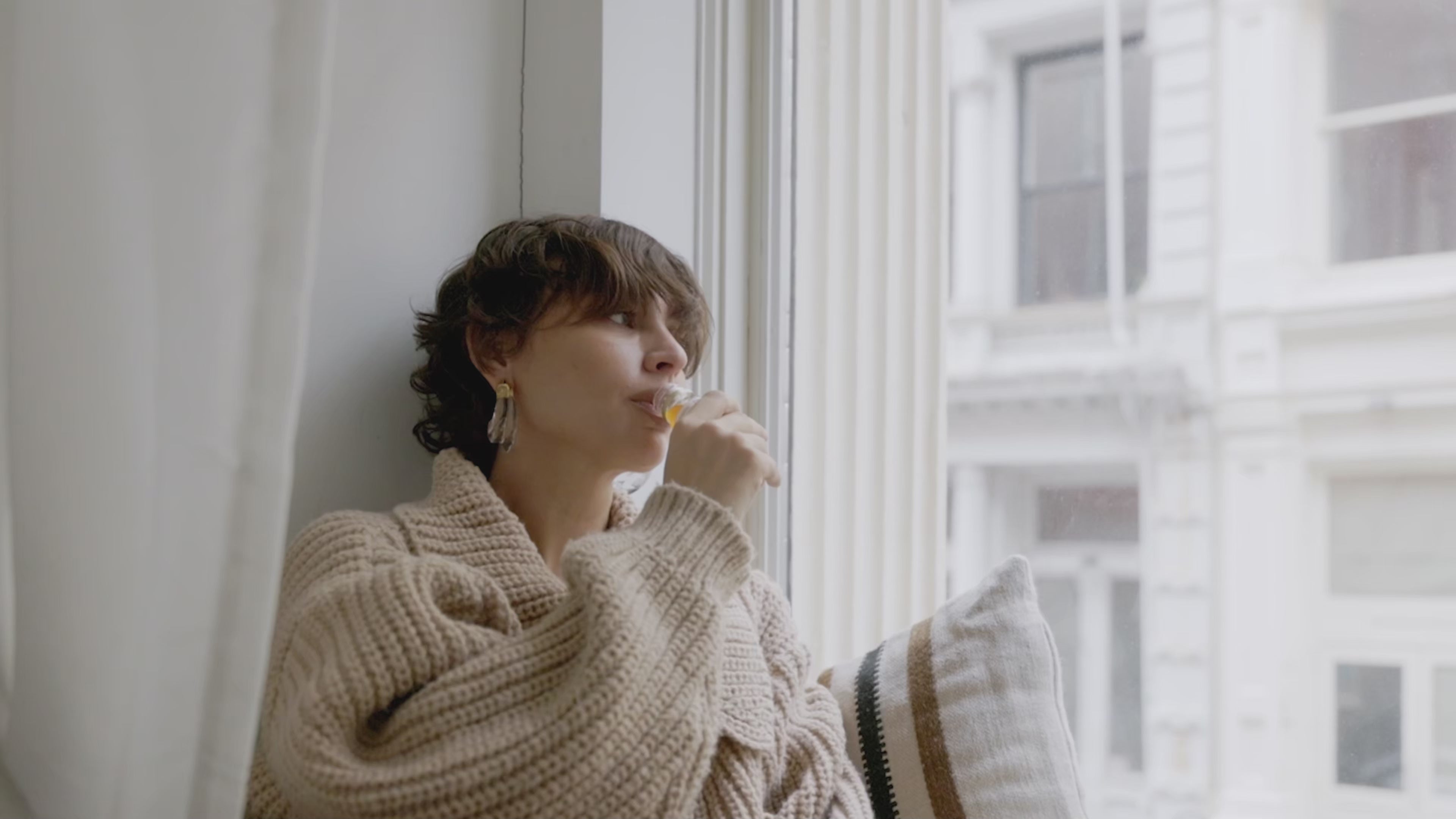 Woman in a cosy sweater drinks Primabiotic Collagen by the window, enjoying a relaxing beauty ritual.