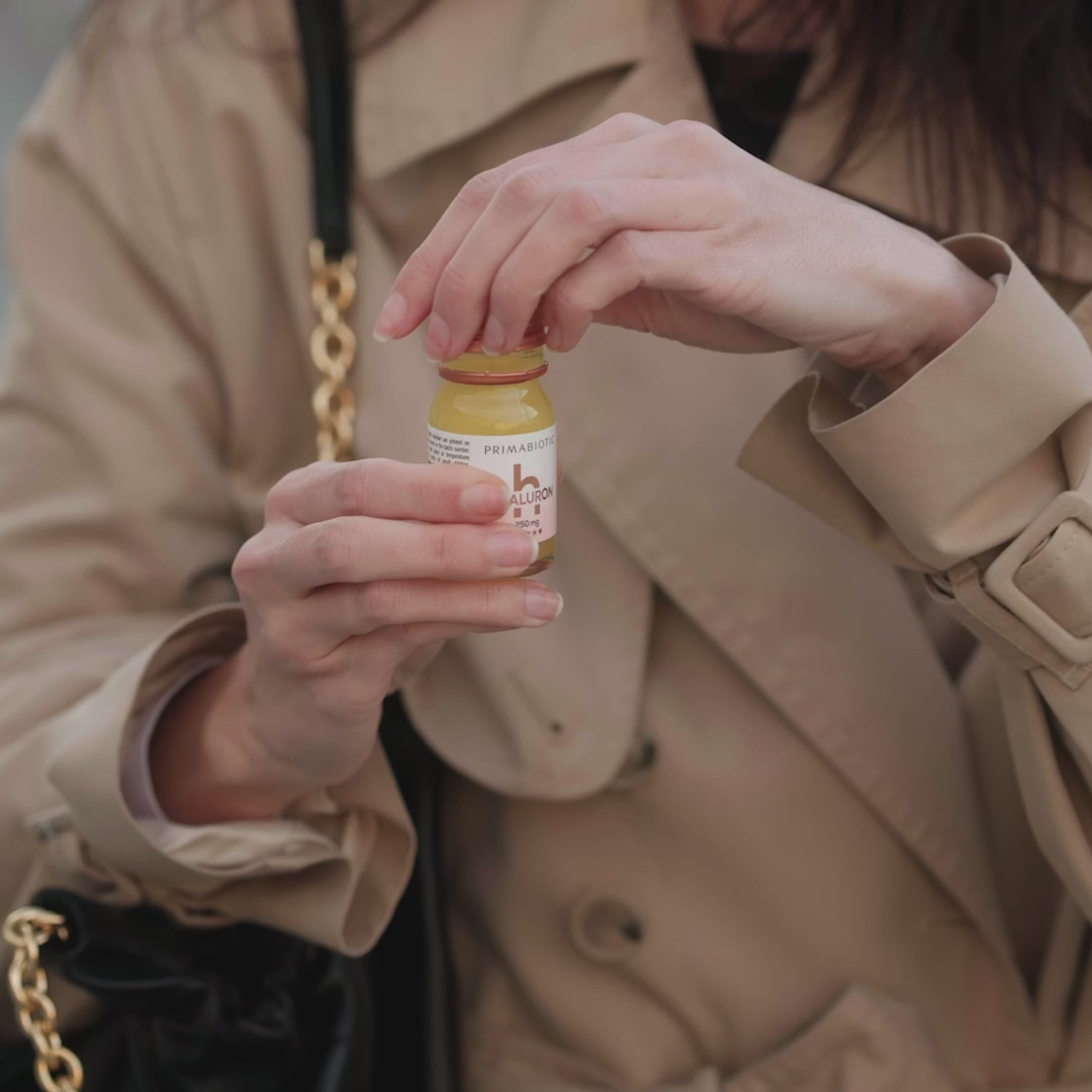 Woman in a beige trench coat holding a small Primabiotic Hyaluron bottle, preparing to open it outdoors.
